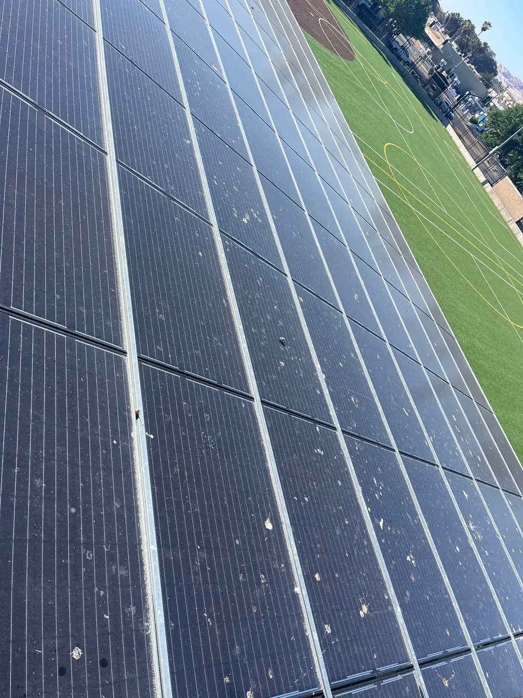 Solar carport modules covered in bird droppings and dust before cleaning, athletic field visible beyond the canopy, school district site, Pittsburg CA